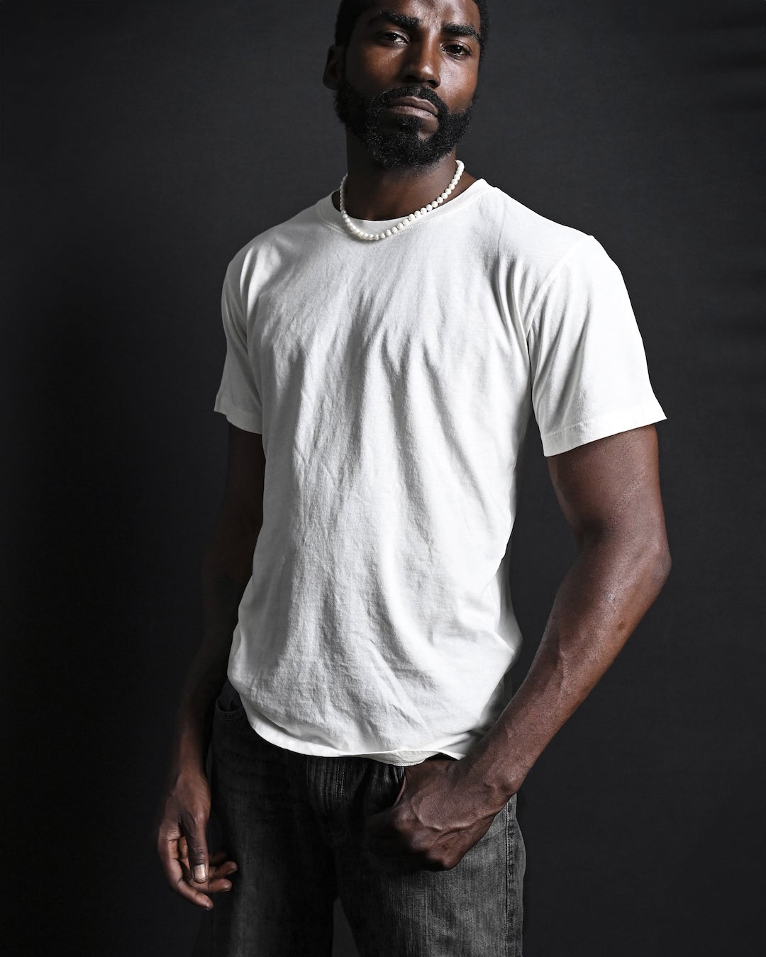 Man in a white t-shirt and necklace, standing against a dark background.